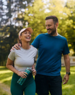 young man and woman walking in nature, smiling, the man has his right arm wrapped around the woman's shoulders, she is holding a water bottle